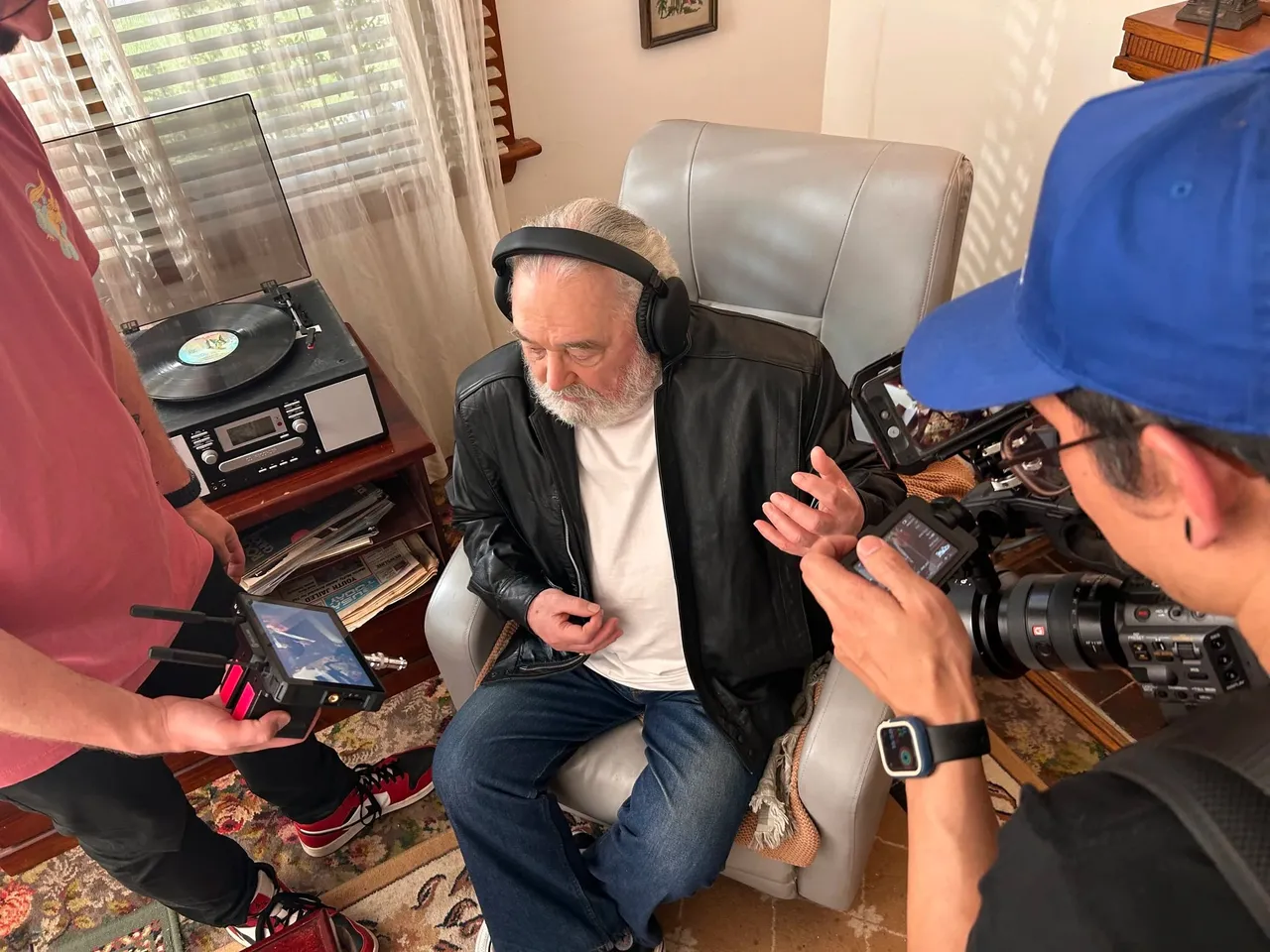 An older man in headphones being filmed in his armchair at home, beside a record player