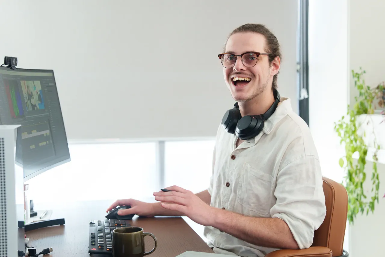 A smiling editor in glasses, headphones round the neck, works at an editing desk with the edit on a monitor alongside.