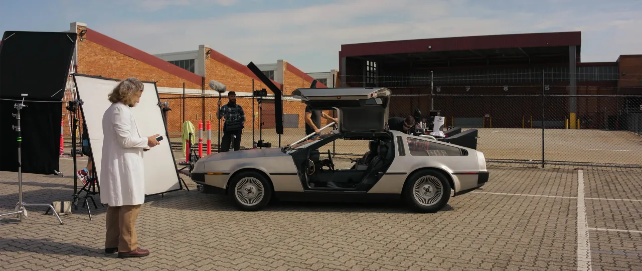 An actor in a white lab coat and grey wig stands beside a DeLorean with its doors raised, on a set with lights and crew in a car park.