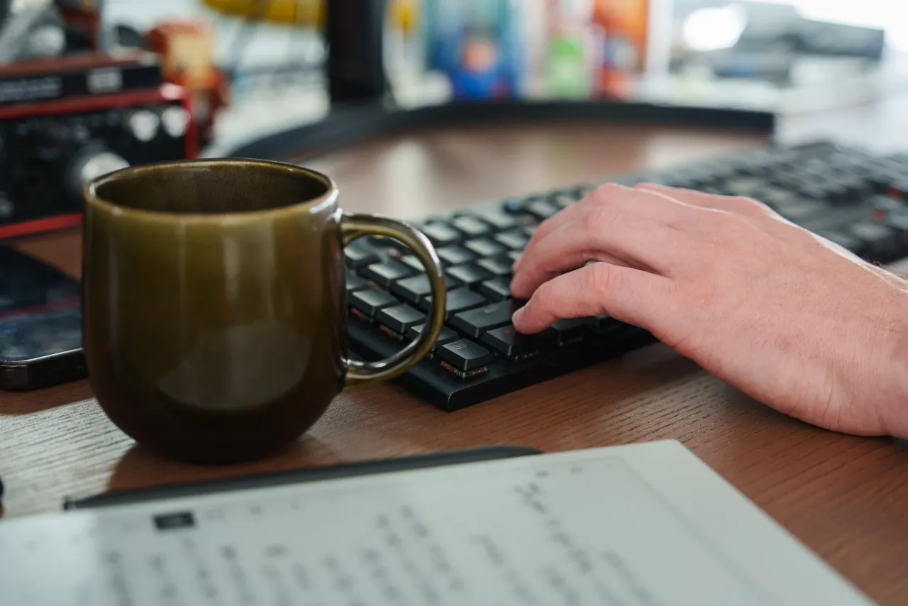 A hand on a keyboard beside a coffee mug and a printed script