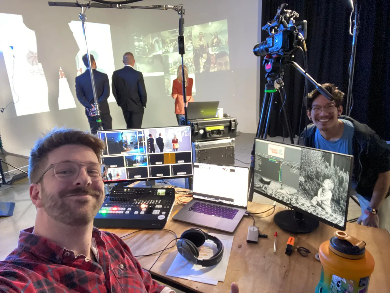 A smiling crew member at a live-streaming desk with a vision switcher and monitors, presenters in the background
