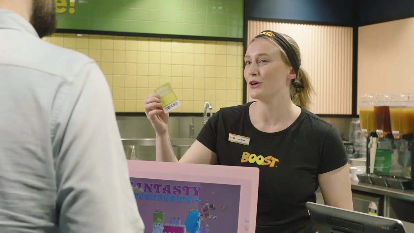 A Boost employee behind the counter, with a black Boost t-shirt and a name tag reading 'Alisha,' engaging with a customer at a smoothie or juice shop. The store has colorful tiled walls and beverage dispensers in the background.