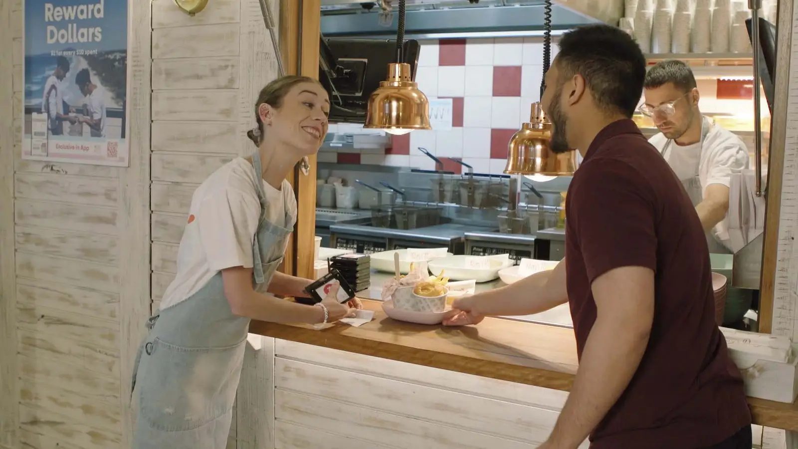 A woman at a restaurant counter receiving food from a man. There are two workers in the background, one wearing glasses and a white apron. The setting appears to be a casual eatery with a white rustic decor and copper pendant lights.