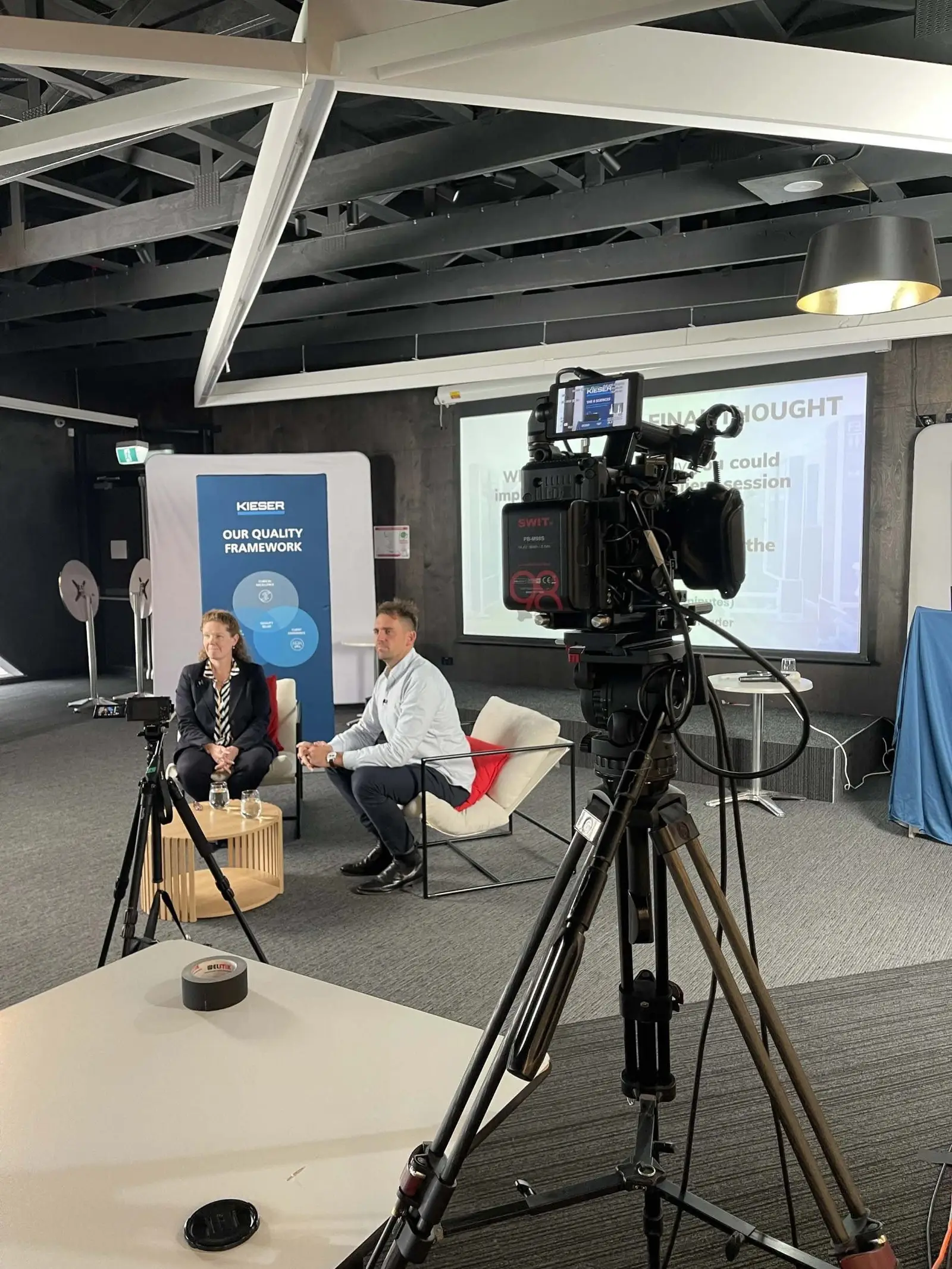 A camera on a tripod set up in front of a woman and a man sitting on a couch, with a presentation screen and a blue poster in the background, in an indoor conference room.
