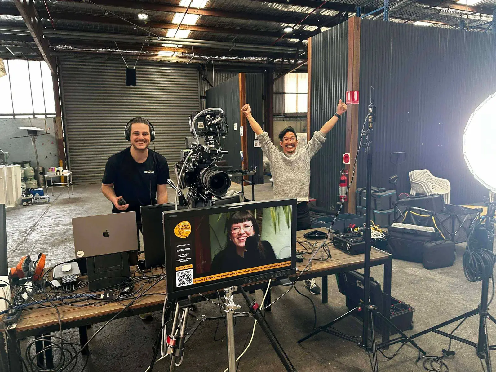 Two men in a warehouse or workshop, one with headphones and camera equipment, the other enthusiastically raising both arms. In front of them, there is a computer monitor and filming equipment, including a camera, wires, and lighting setup. The background has metal walls, a closed roller door, and shelves with various items.